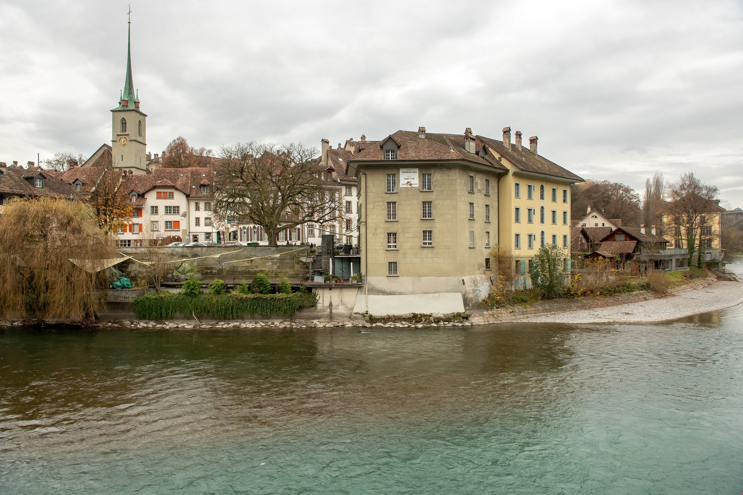 A building with a clock tower, situated on the bank of a river, surrounded by other buildings and trees.