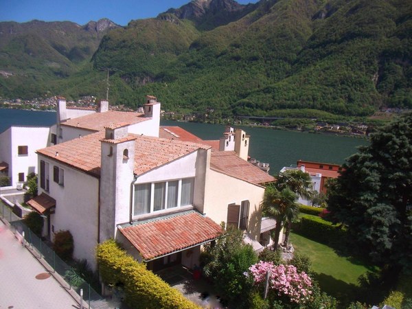 Large house with red roof, white walls, multiple windows, surrounded by greenery, mountains in the background, lake nearby