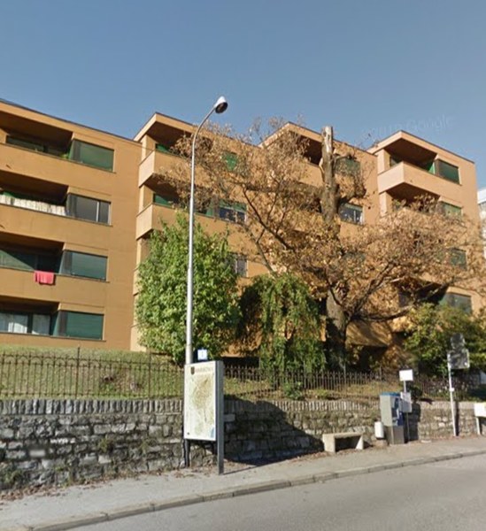 Apartment building with 4 stories, large windows, concrete fence, paved sidewalk, trees