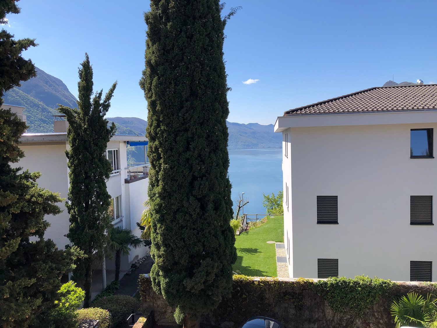 Two white buildings, one with a balcony, surrounded by greenery, with a lake and mountains in the background.