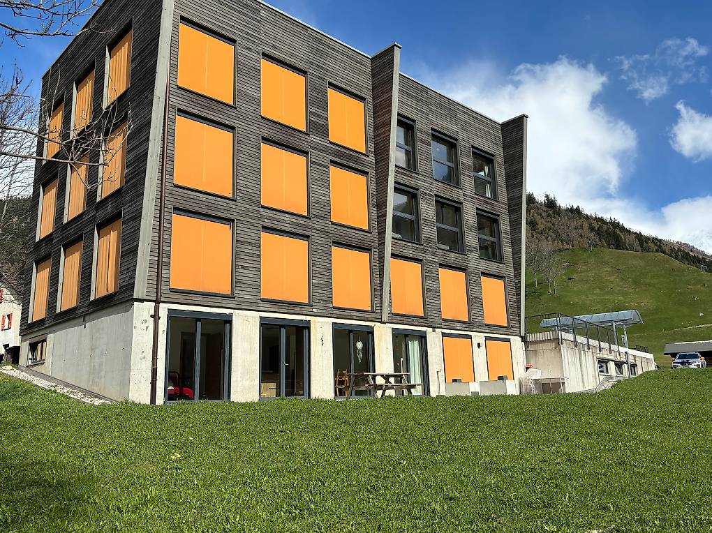 A modern building with gray and orange exterior, raised ground floor, large glass windows, surrounded by green grass and a car parked outside.
