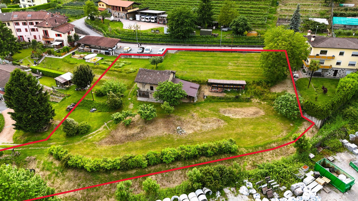 Empty plot of land with house in the middle, red line indicating boundary, surrounding are houses, cars parked, street light, trees, vineyard
