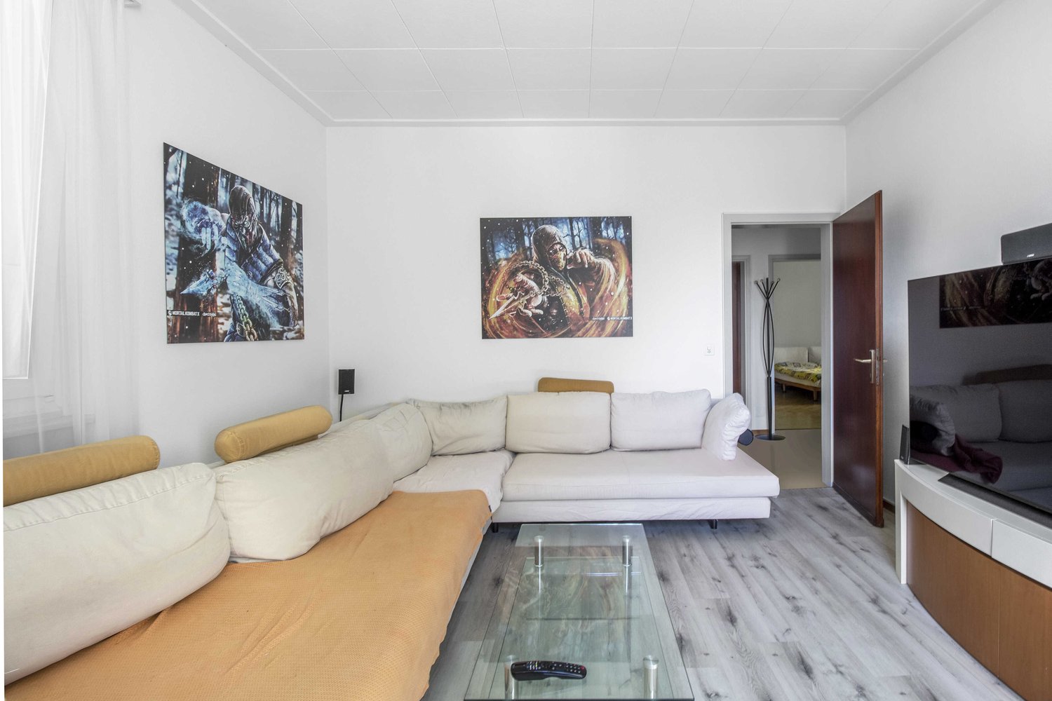 L-shaped sofa, glass coffee table, two paintings on the wall, TV, remote control on the table, white curtains