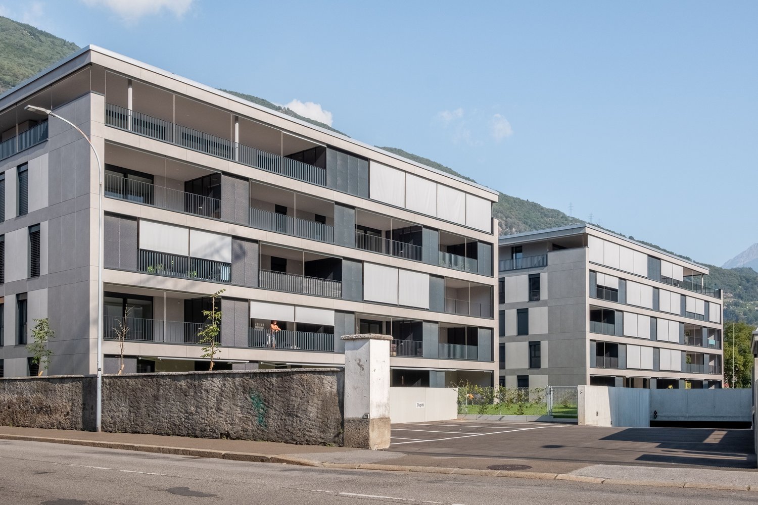 Gray exterior, large windows, balconies, street light, stone wall, underground parking, grass, mountain in the background