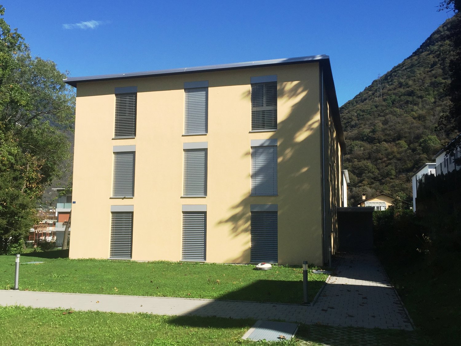 Modern yellow building with 8 windows and a flat roof, with grass and trees around, located on a raised ground floor