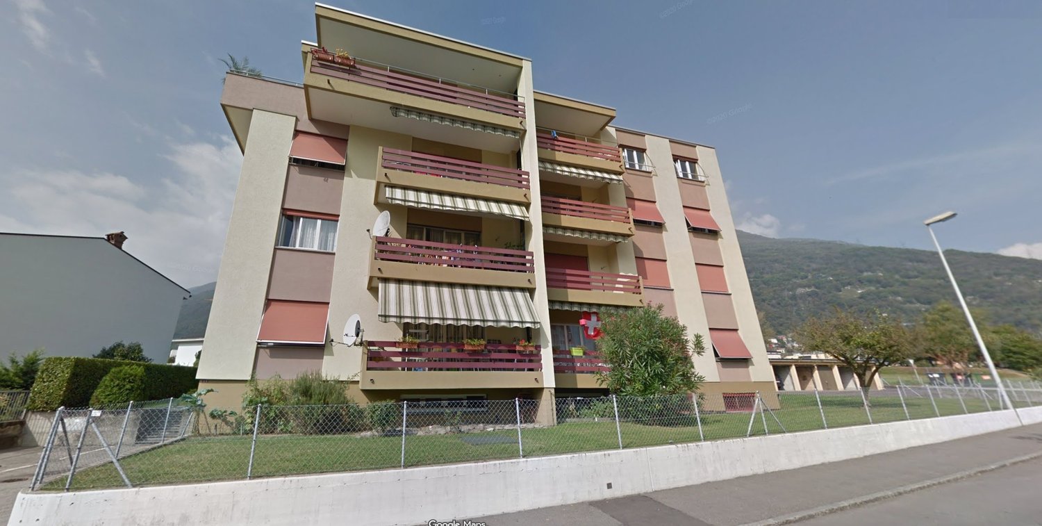 Multi-story apartment building with multiple balconies, red and beige walls, surrounded by grass, a streetlight and a fence.