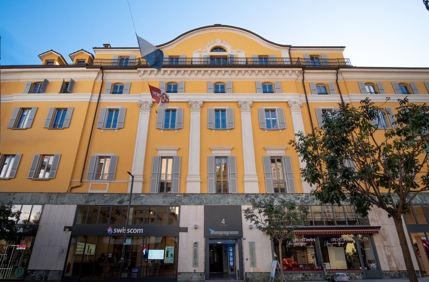 Yellow building with multiple windows, flags, entrance, and shops on the ground floor