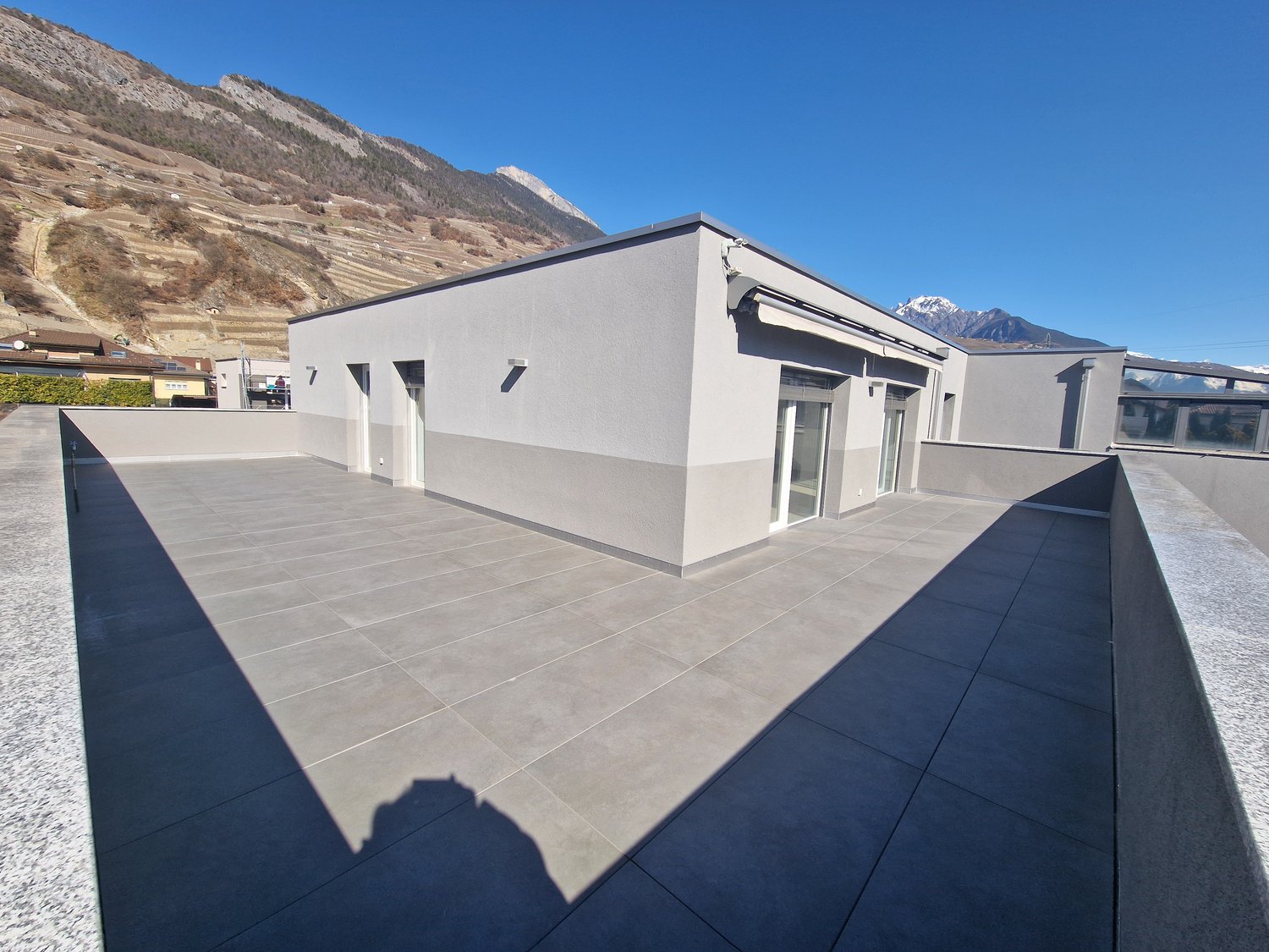 A modern, two-story building with a flat roof and large windows. The exterior is painted in a light gray color. The building has a spacious terrace or balcony area with a tiled floor. The surrounding landscape includes mountains in the background.