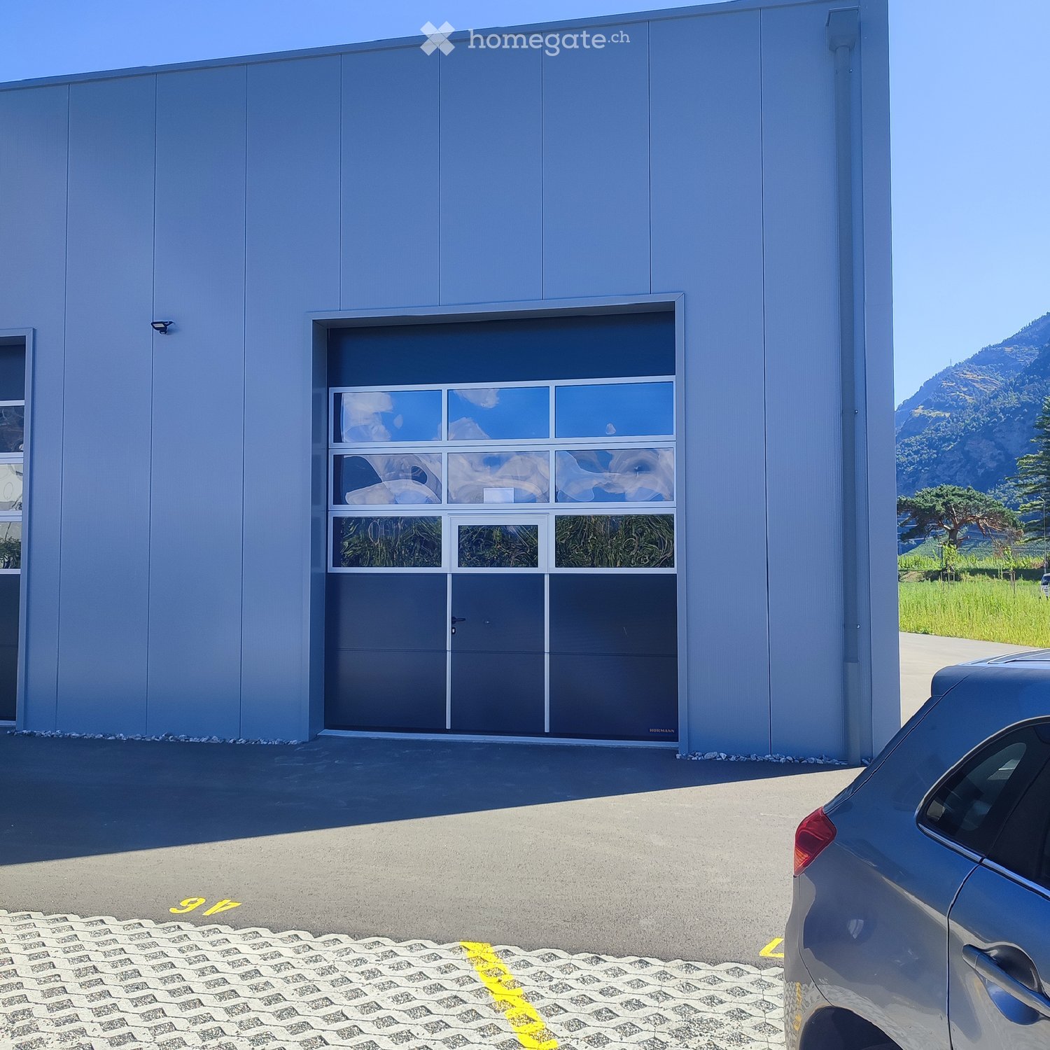 Industrial building, grey exterior, large roll-up door, glass panels, surveillance camera, paved area, yellow markings, grass field, trees, mountain in distance