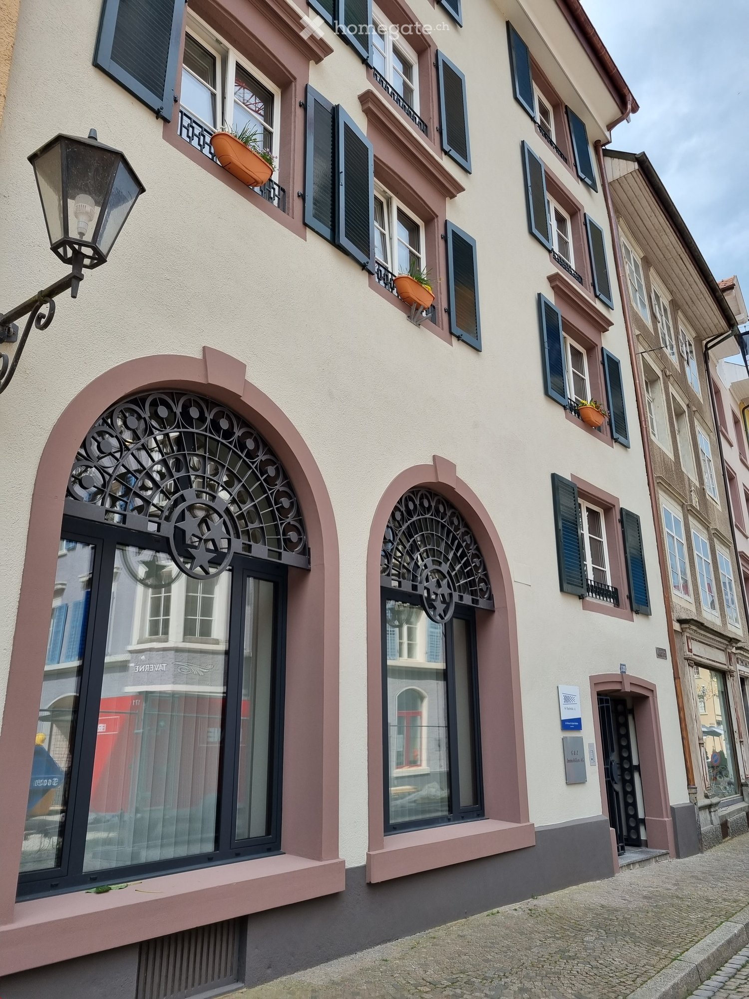 Urban facade, beige walls, three arched windows with black metal grilles, three balconies with potted plants, two lamps attached to the wall