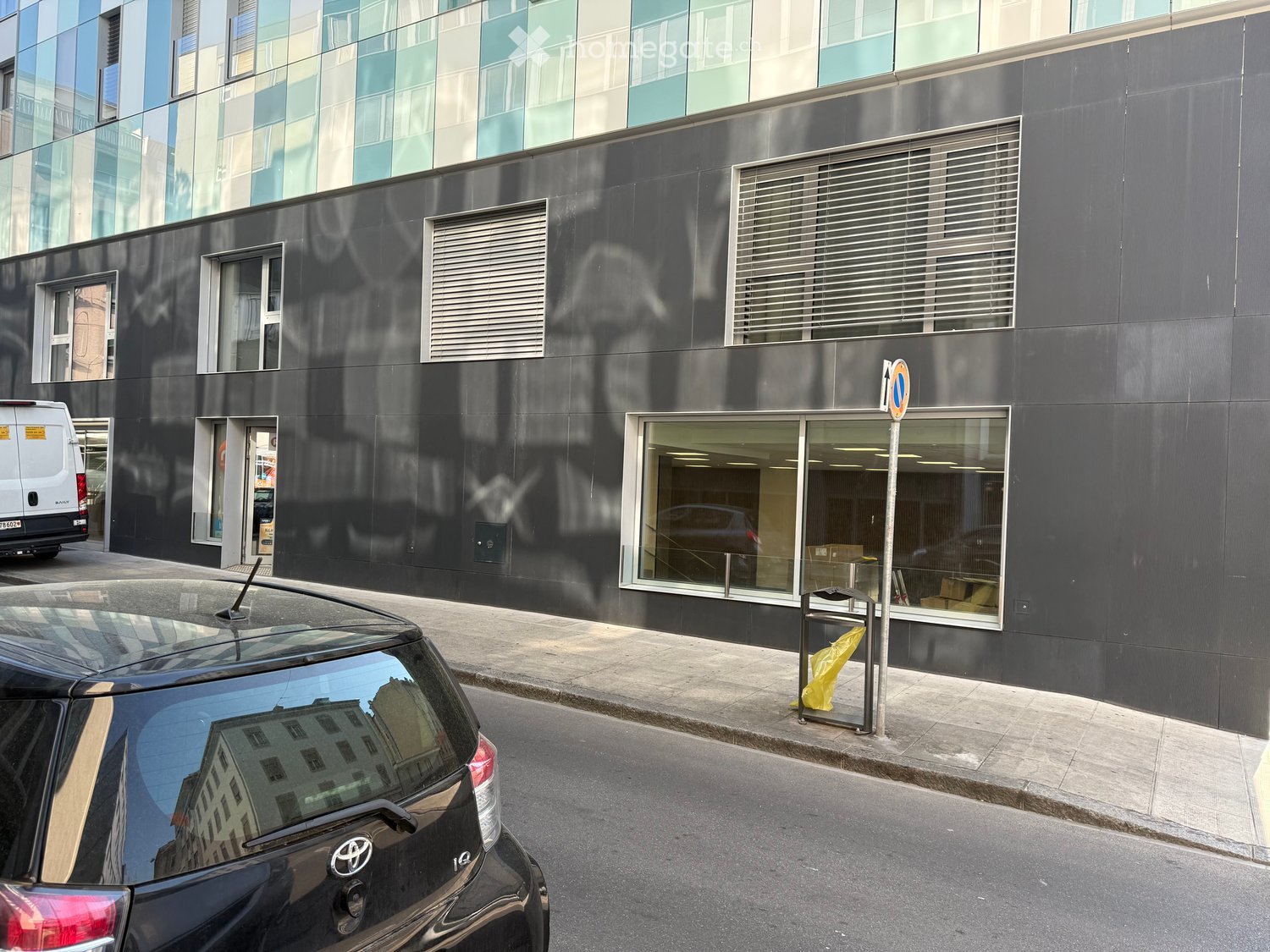 Modern building with glass facade, multiple windows, black exterior, glass door, trash bin, parked car