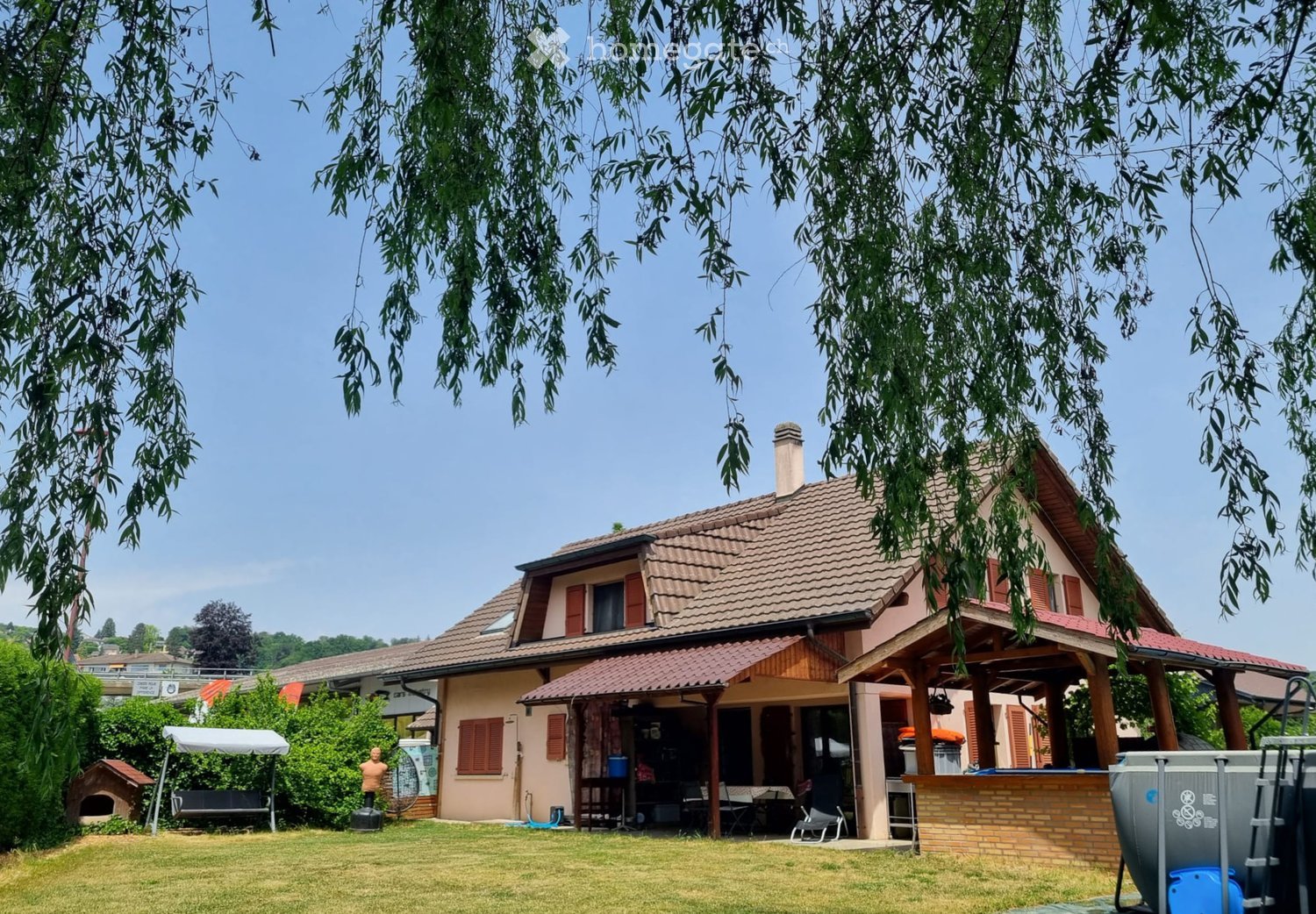 2 story house, brown roof, brick wall, wooden covered area, swimming pool, large lawn, gazebo, trees, bushes, dog house