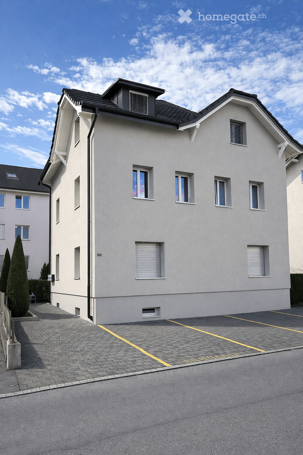 Two story house with beige walls and black roof, multiple windows, two parking places in front