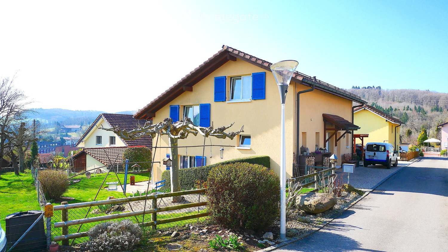 House. Two-story house with yellow walls, blue shutters, and a tiled roof. The house has a well-maintained garden with a wooden fence, a tree, and a small bench. A white van is parked in the driveway.