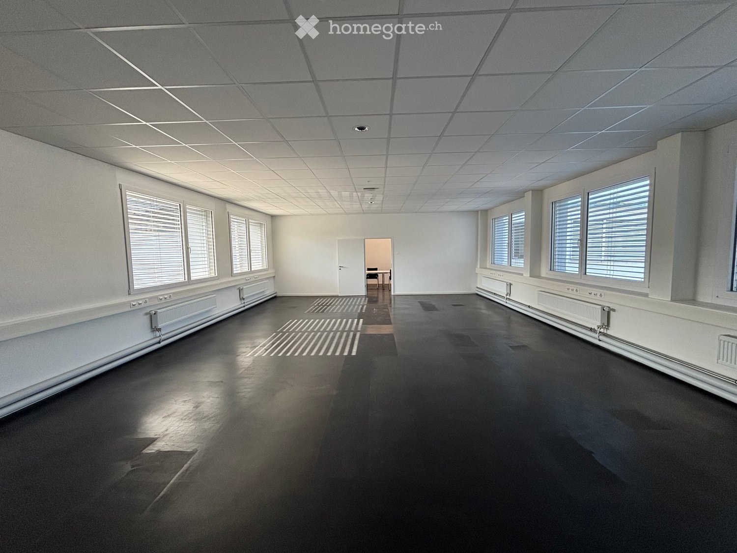 Empty room with white walls, black floor, large windows with blinds, and a doorway leading to another room.