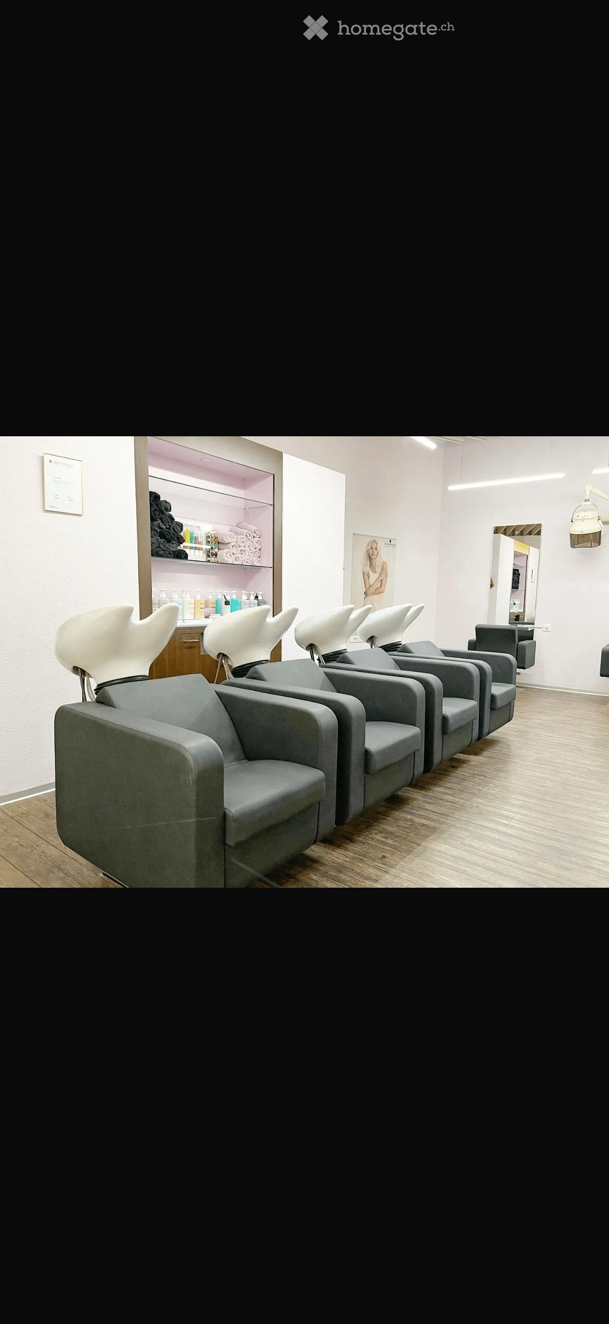 A salon with a row of grey chairs, a wooden floor, a white wall, a shelf with products, and a mirror.