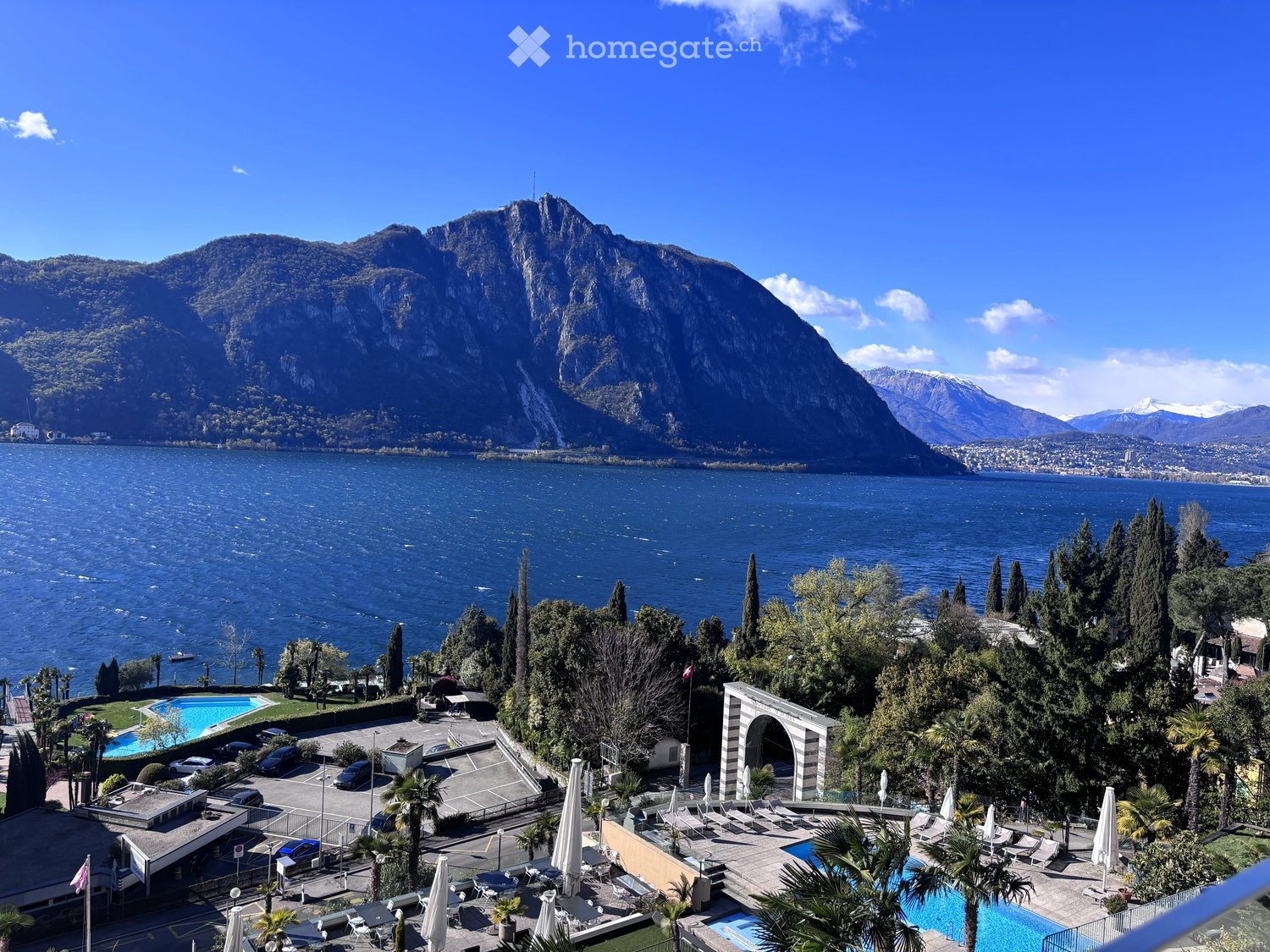 Aerial view of a resort with a swimming pool, mountains, and a lake in the background
