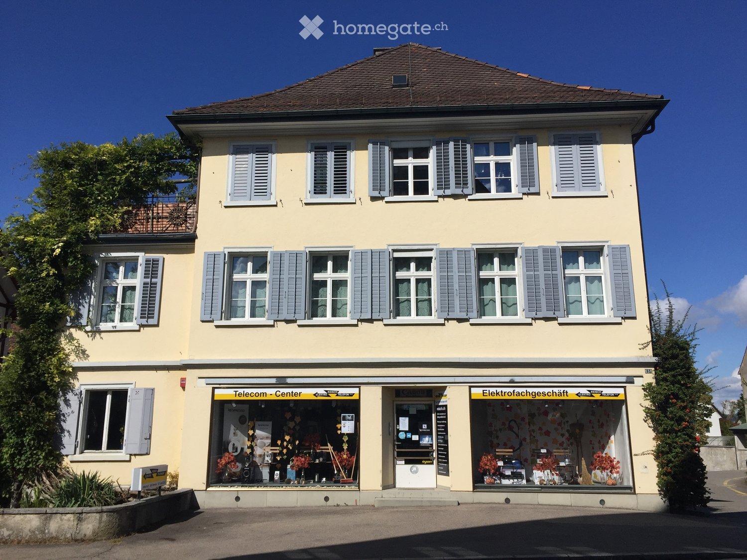 3 story house, yellow exterior, brown roof, multiple windows, balcony, storefronts