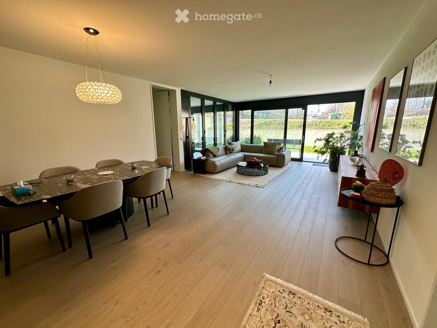 Dining room with wooden floor, large windows, dining table, chairs, couch, coffee table, rug, plants, lamp