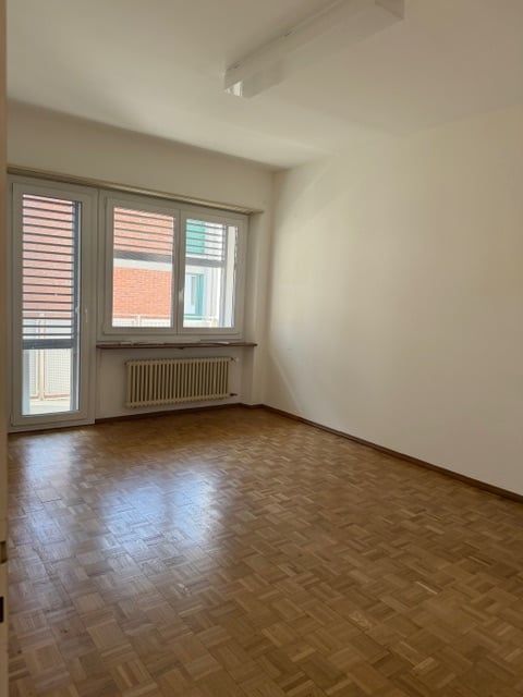 Empty room with wooden floor, white walls, large windows, sliding glass door, and a radiator.