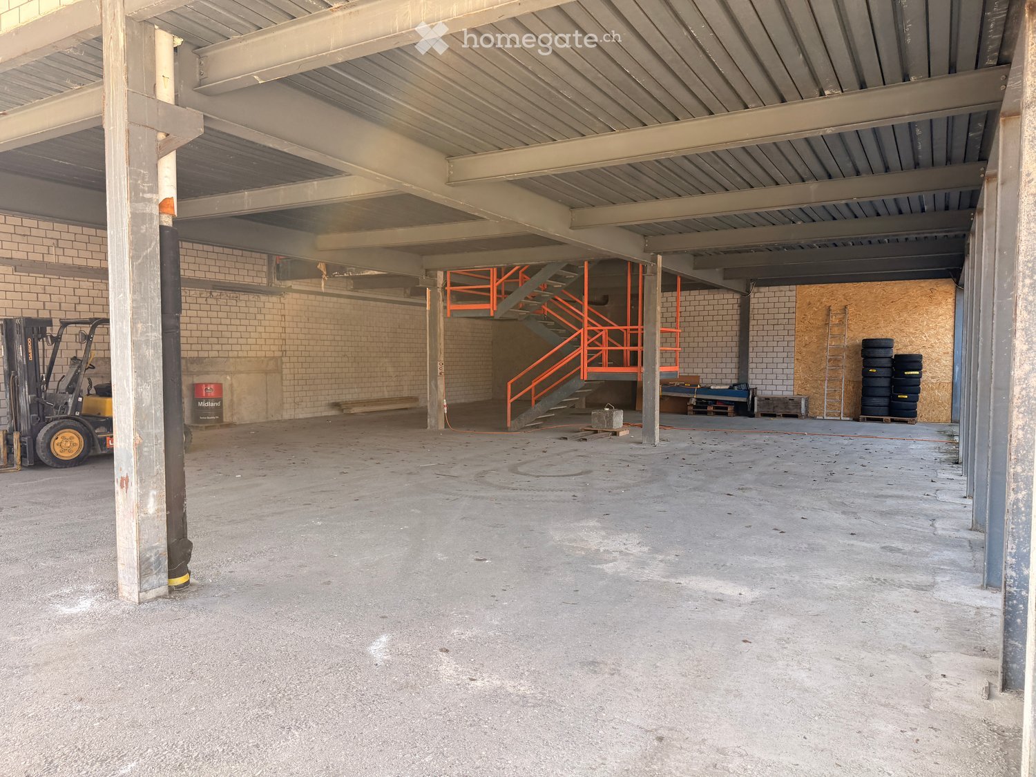 Empty warehouse with metal beams, brick walls, concrete floor, orange staircase, forklift, tires, ladder