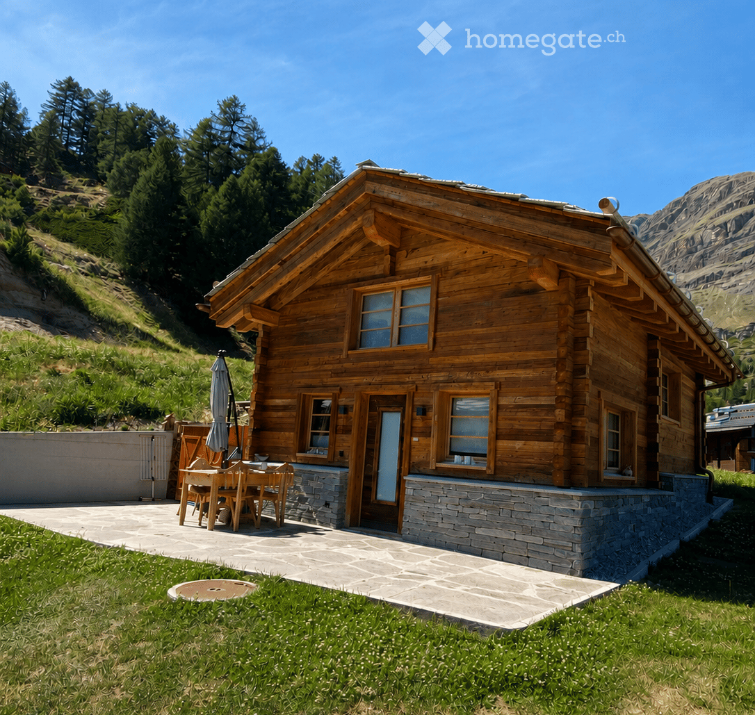 A wooden chalet-style house with a stone base, large windows, and a raised ground floor. The house is surrounded by a grassy area and a stone patio with outdoor furniture.