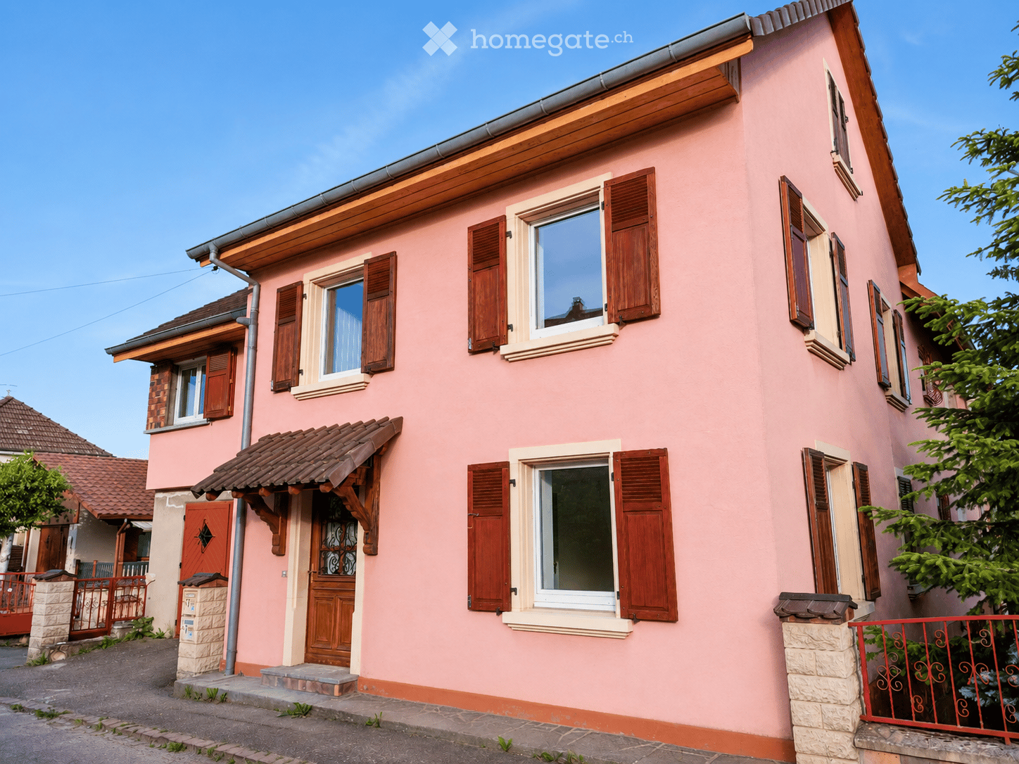 Two-story pink house with wooden shutters, multiple windows, and a tiled roof. Features a small porch with a wooden door and a stone base.