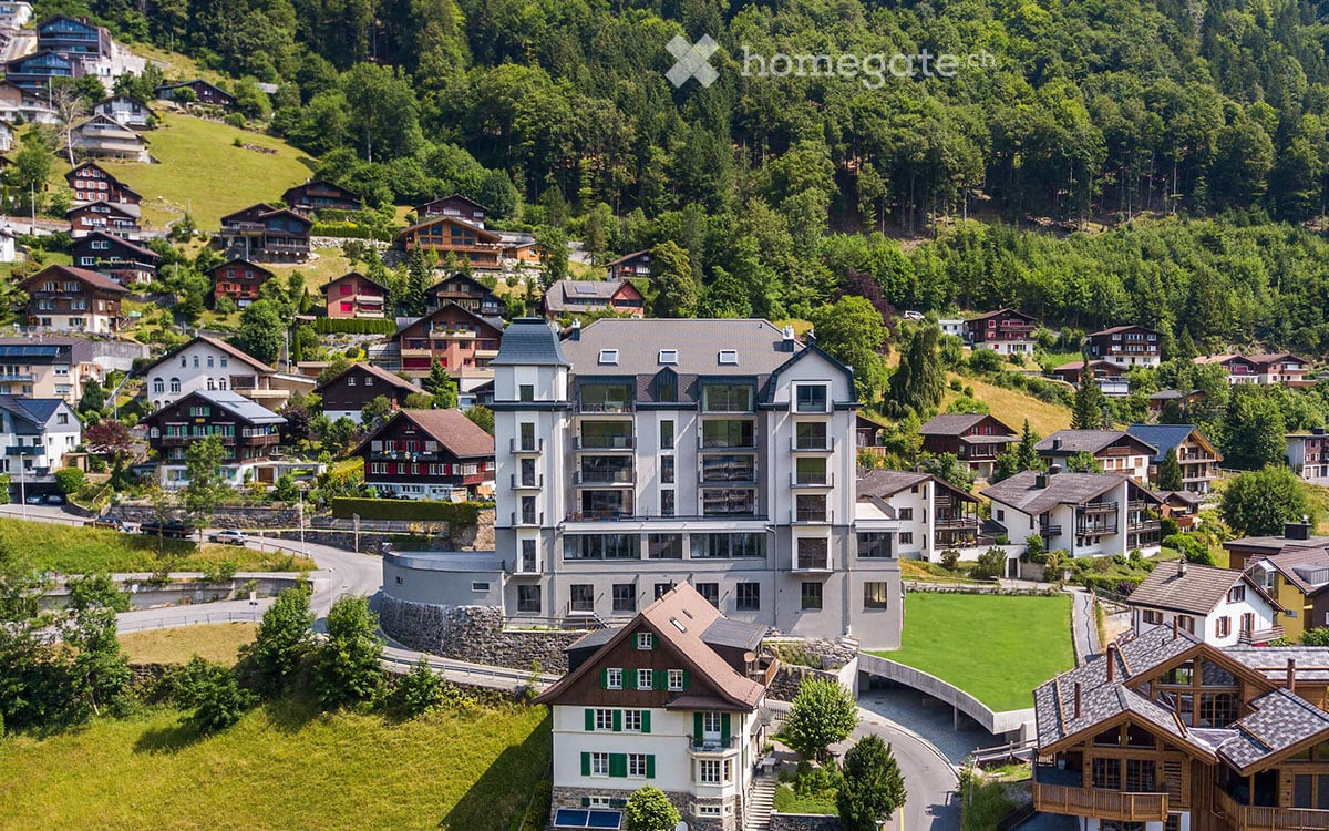 Aerial view of a modern apartment building surrounded by houses and trees, set on a hillside with a scenic valley below.