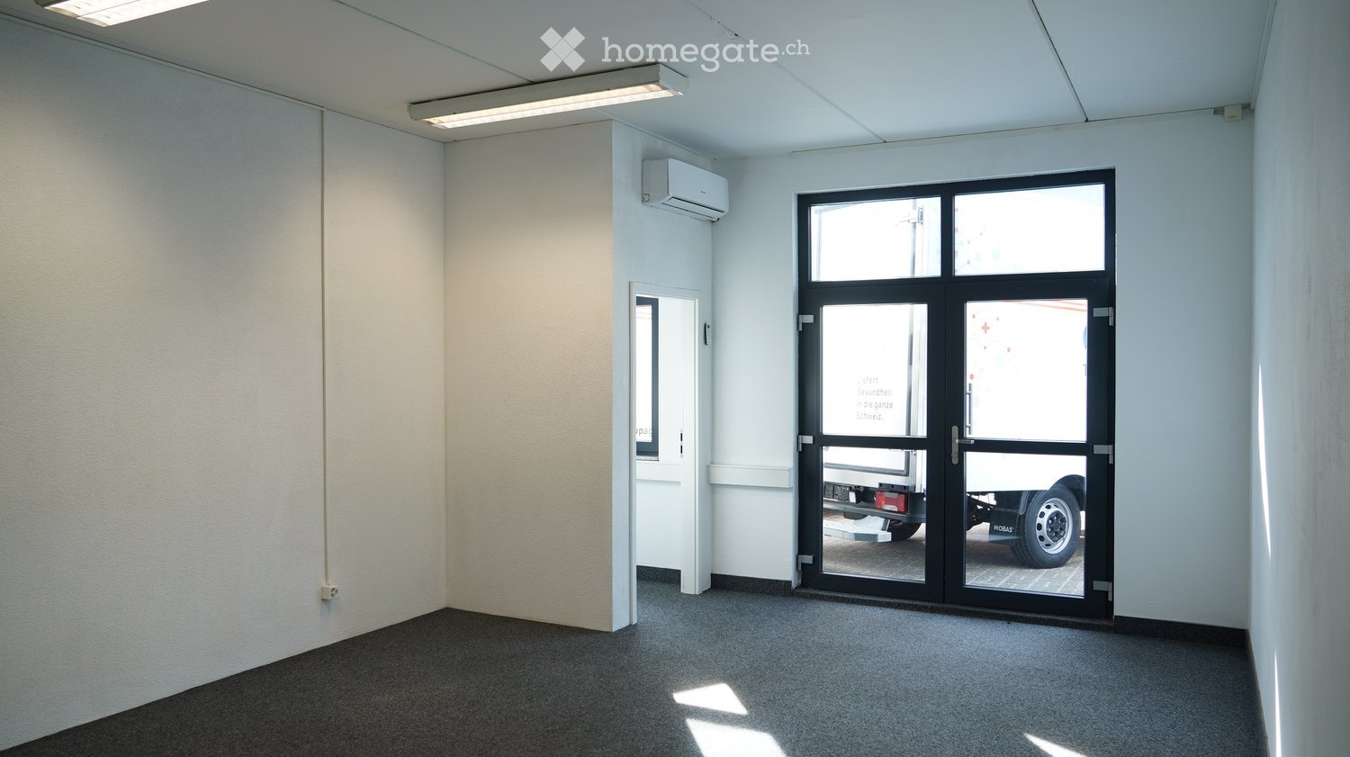 Empty room with white walls, gray carpet, and a glass door. There is a small window on the left wall and an air conditioning unit mounted on the wall above the door.
