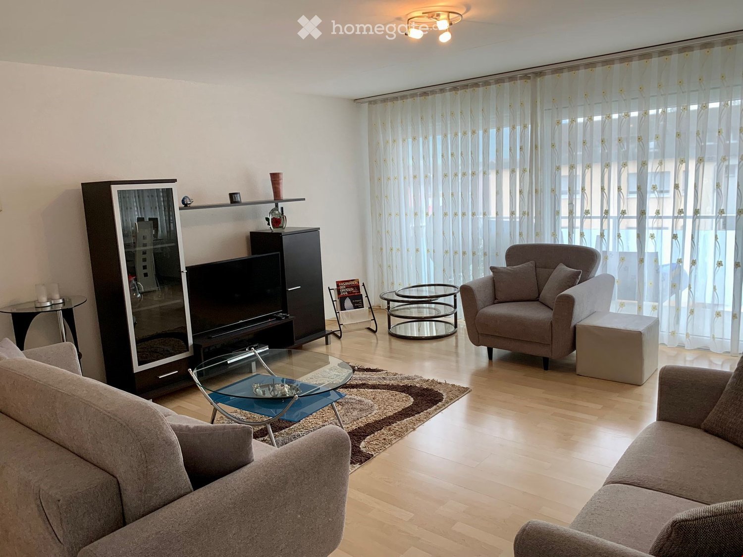 Spacious living room with light wood floors, large windows, and white curtains. Features a central coffee table, a rug, and a TV on a cabinet. Two sofas with pillows and a chair with a side table. A book rack and candles add to the decor.