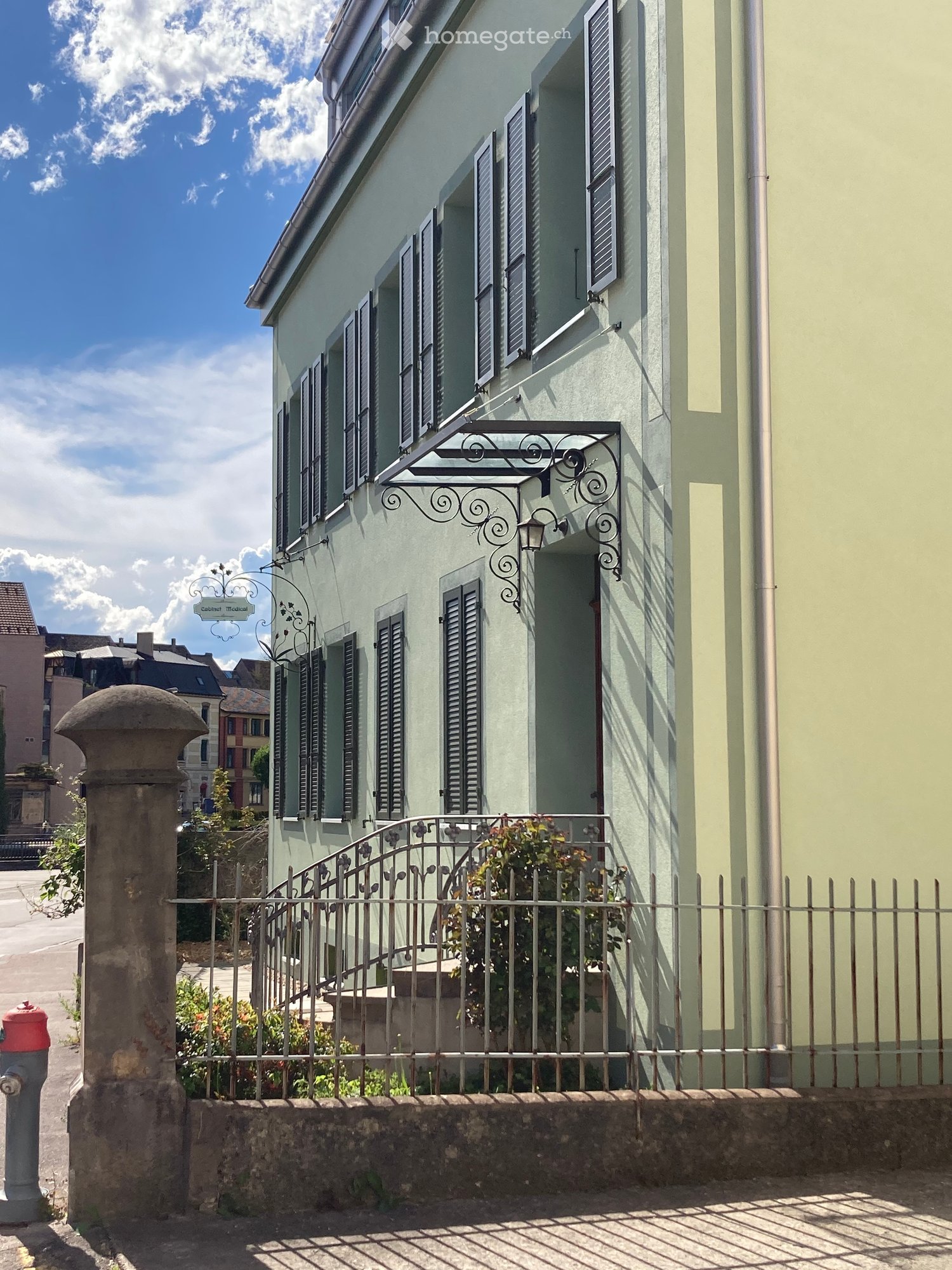 Building. A two-story building with light green walls, multiple windows with shutters, a decorative wrought iron balcony, and a small garden area behind a metal fence.