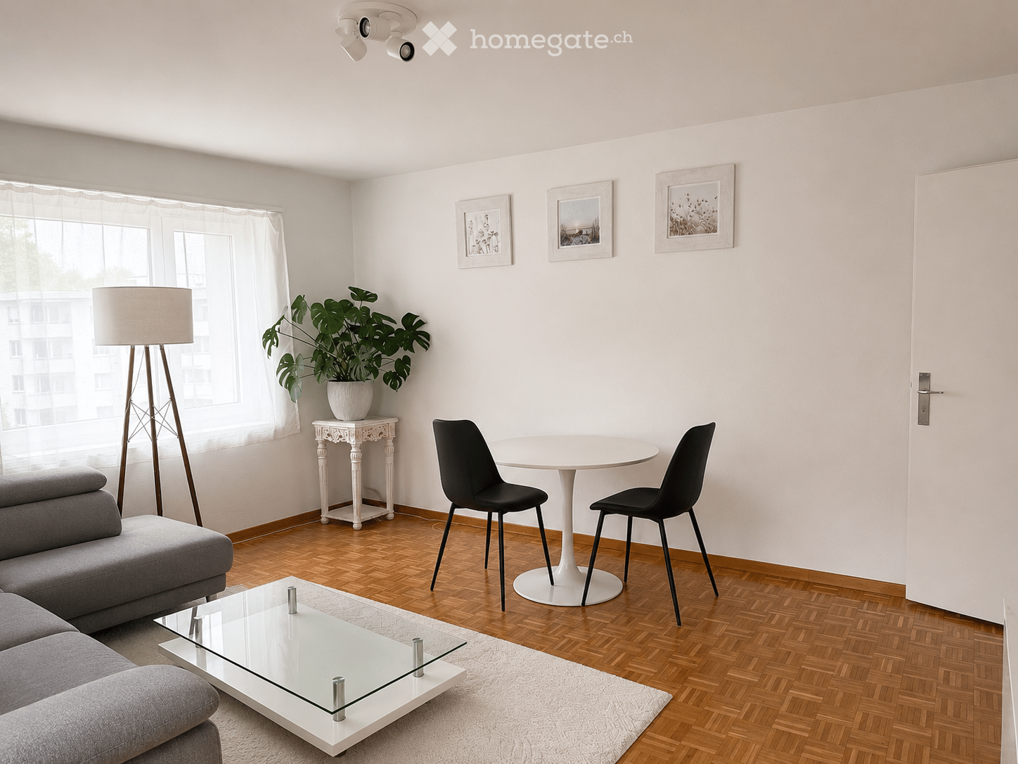 Modern living room with a grey sofa, glass coffee table, white dining table with two black chairs, floor lamp, side table with a plant, and framed artwork on the walls.