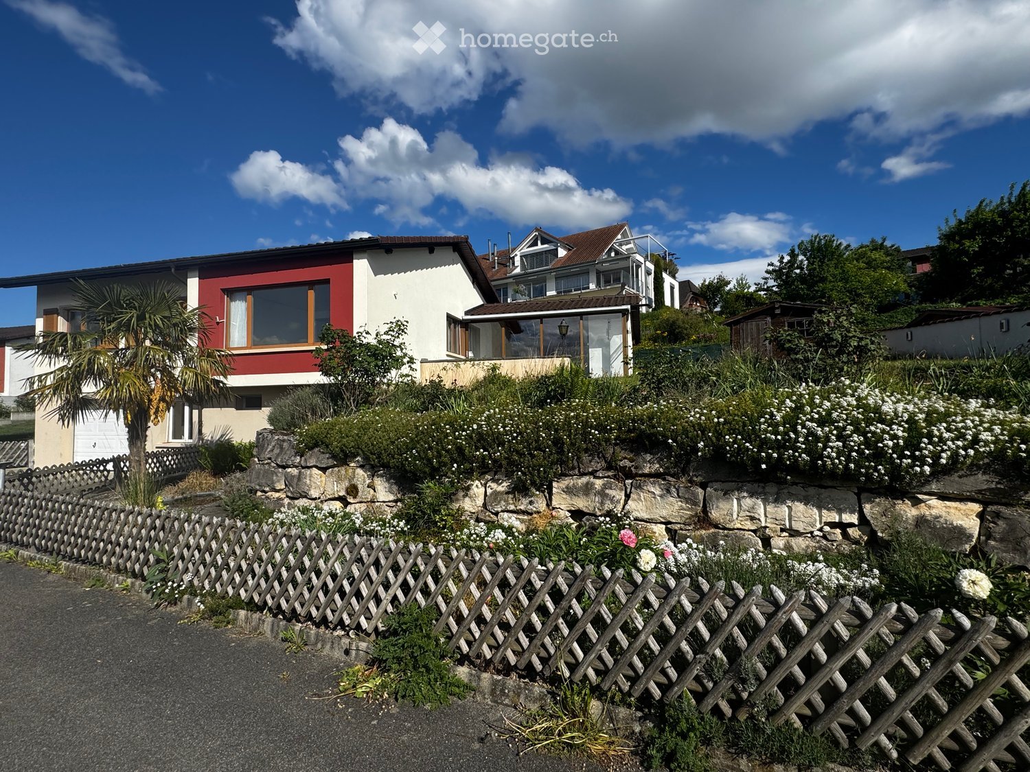 House. Two-story house with red and white exterior, large windows, and a balcony. The house is surrounded by a garden with a wooden fence and various plants.