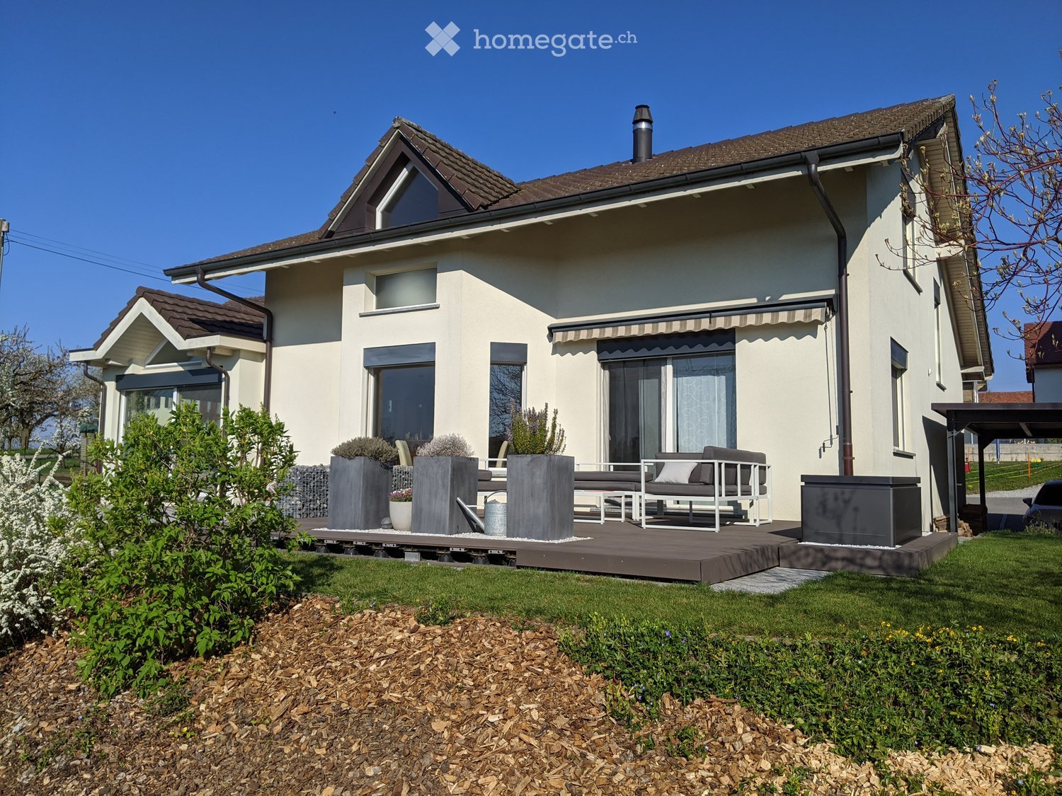 House. Modern two-story house with beige exterior, large windows, and a covered patio area with seating and planters.