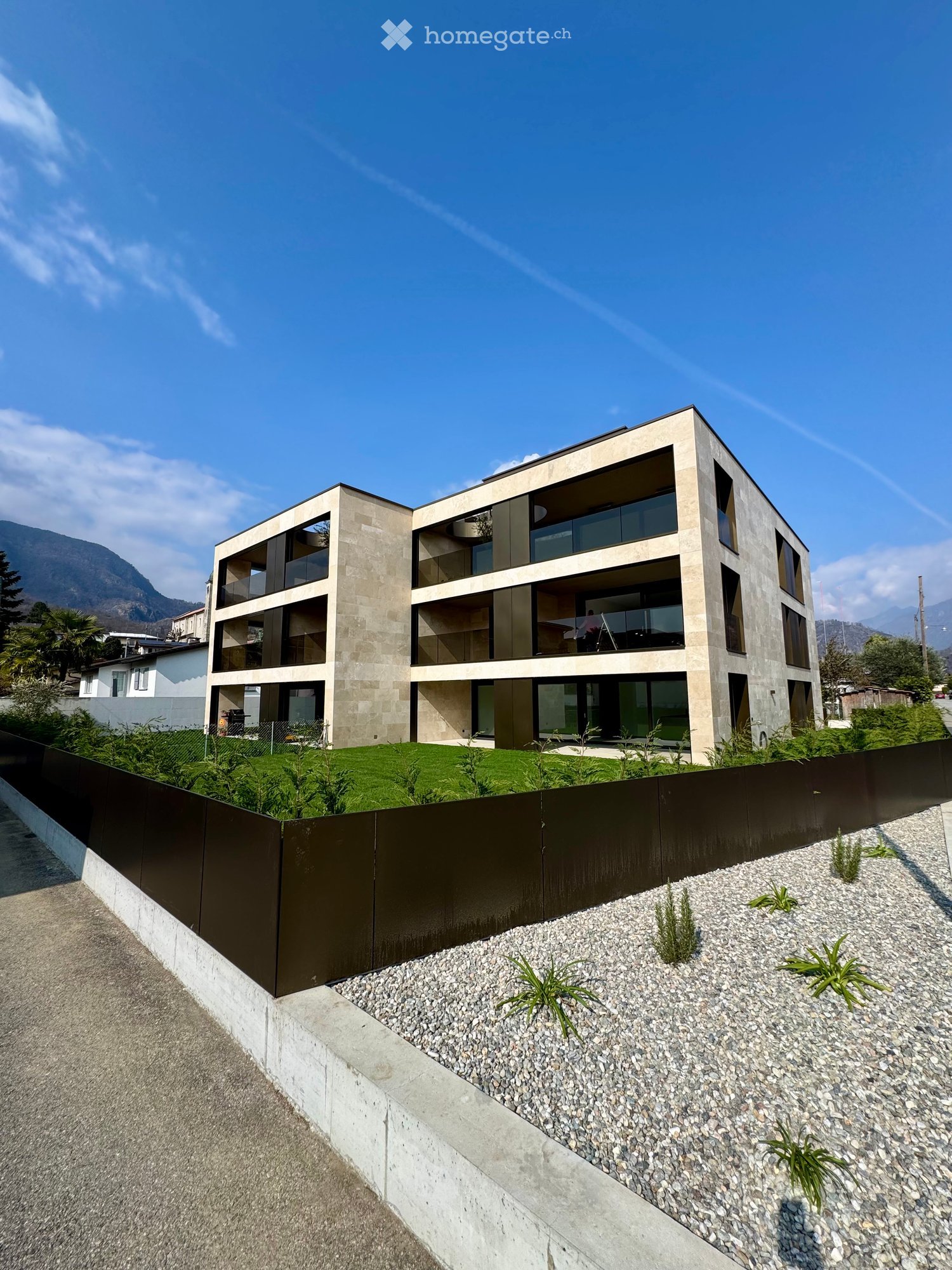 Building. Modern multi-story building with large windows and balconies, surrounded by a low fence and landscaped area with gravel and small plants.