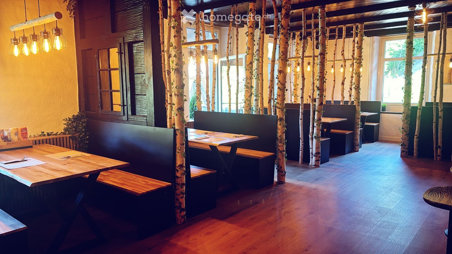 Restaurant interior with wooden tables, benches, and hanging lamps, featuring birch tree trunks as decorative columns.