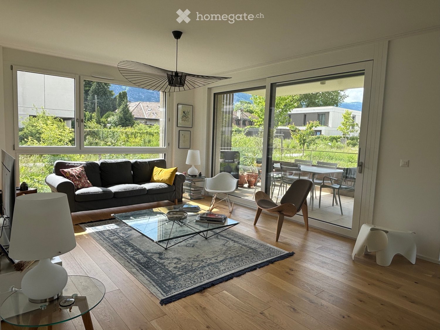 Living Room. Modern living room with large windows and a view, featuring a dark grey sofa, glass coffee table, white lamp, and a dining area with chairs and a table visible through the open sliding doors.