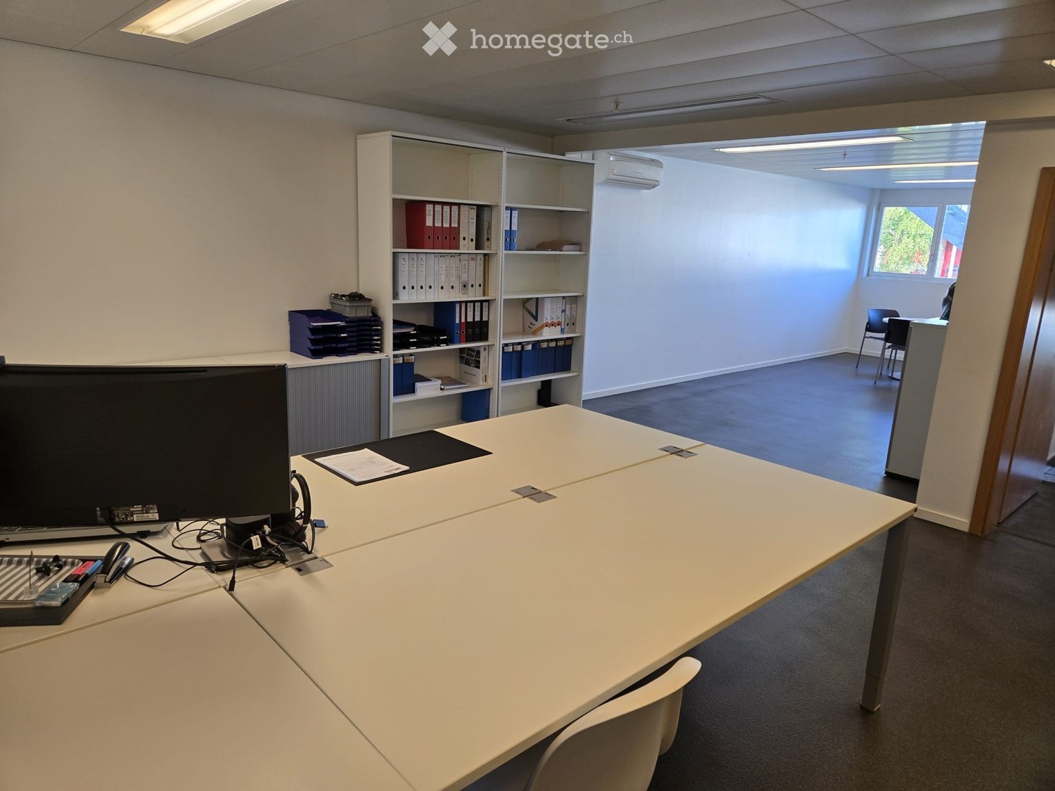 Furnished Room. Office space with a large white table, chairs, a screen, laptop, keyboard, mouse, and shelves with folders and books.