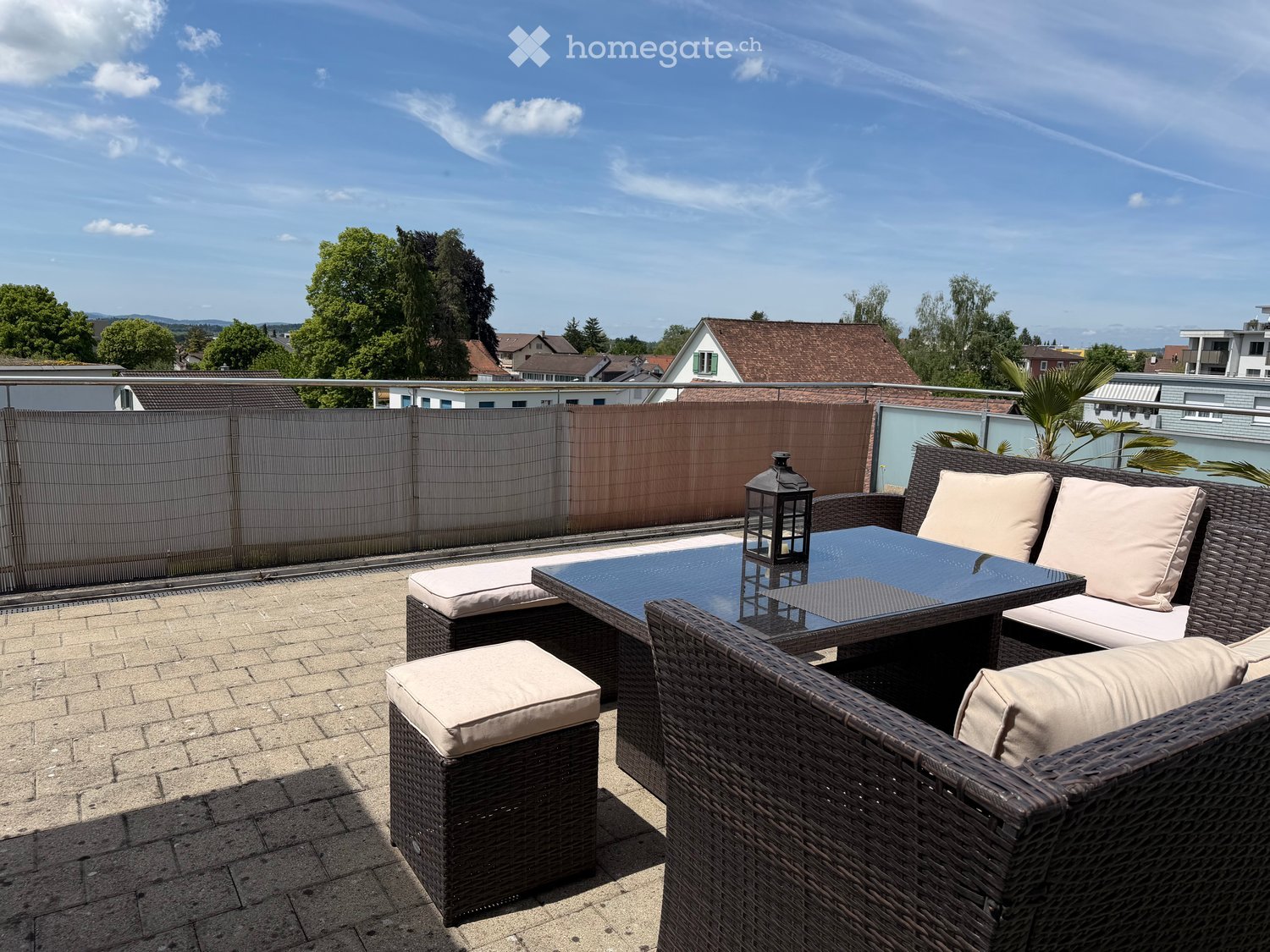 A modern rooftop terrace with a wicker seating set, glass table, and lantern, overlooking a residential area with trees and houses.