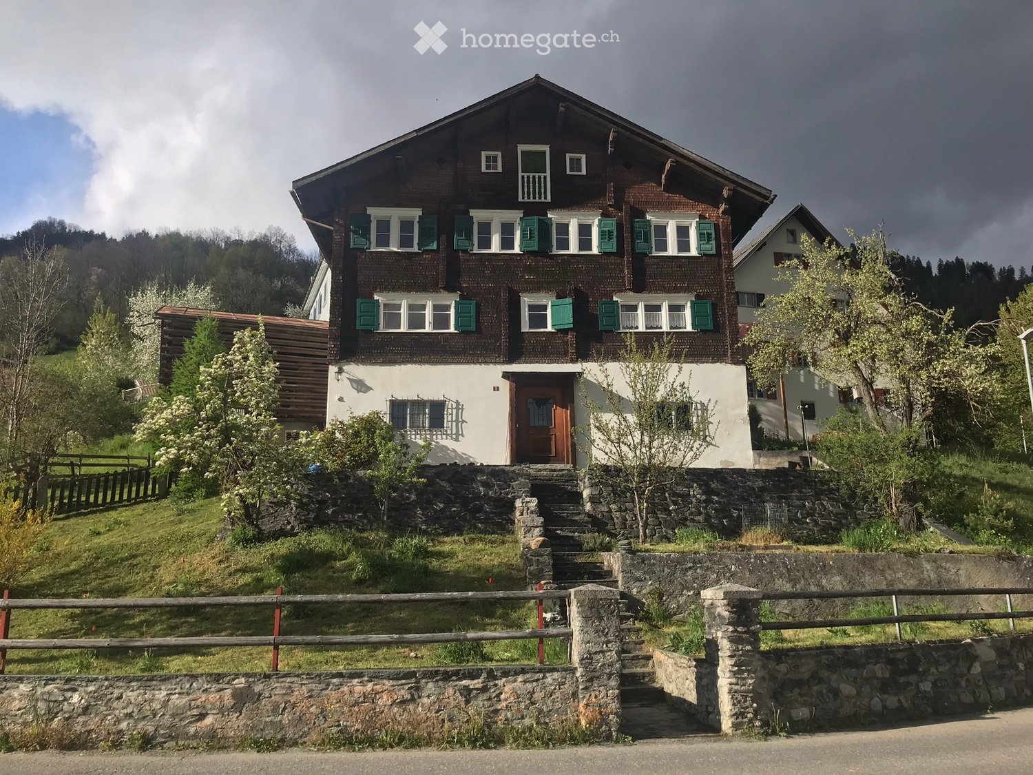 3 story house, brown wooden exterior, green shutters, white walls, stone steps, stone wall, trees, grass, fence