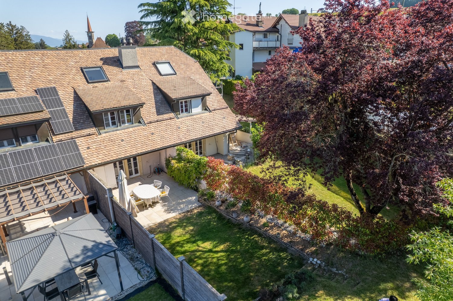 Aerial view of a residential house with a well-maintained garden, patio area with outdoor furniture, and solar panels on the roof.