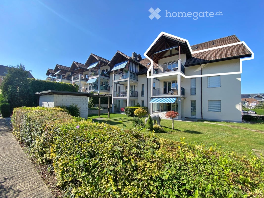 Modern multi-story residential building with balconies, surrounded by well-maintained gardens and hedges.