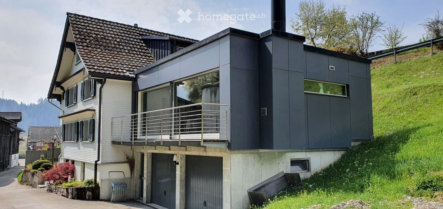 Modern two-story house with a combination of white and dark grey exterior walls, a tiled roof, a raised ground floor, a garage, and a balcony with metal railings.