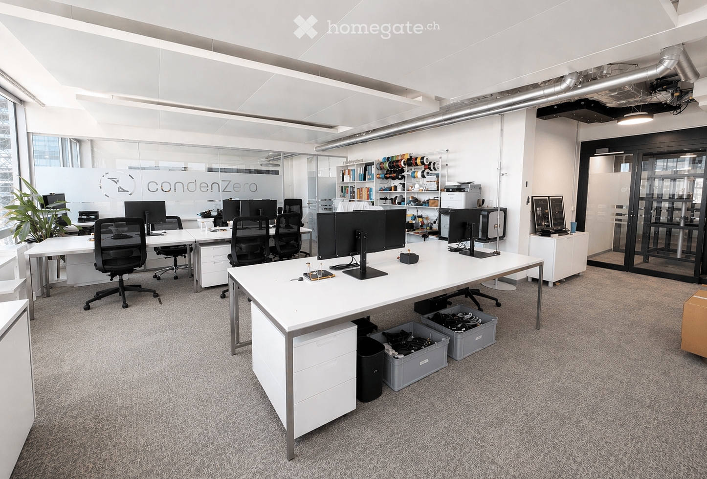 Modern office space with white desks, black office chairs, multiple computer monitors, shelves with office supplies, and a potted plant near the window.