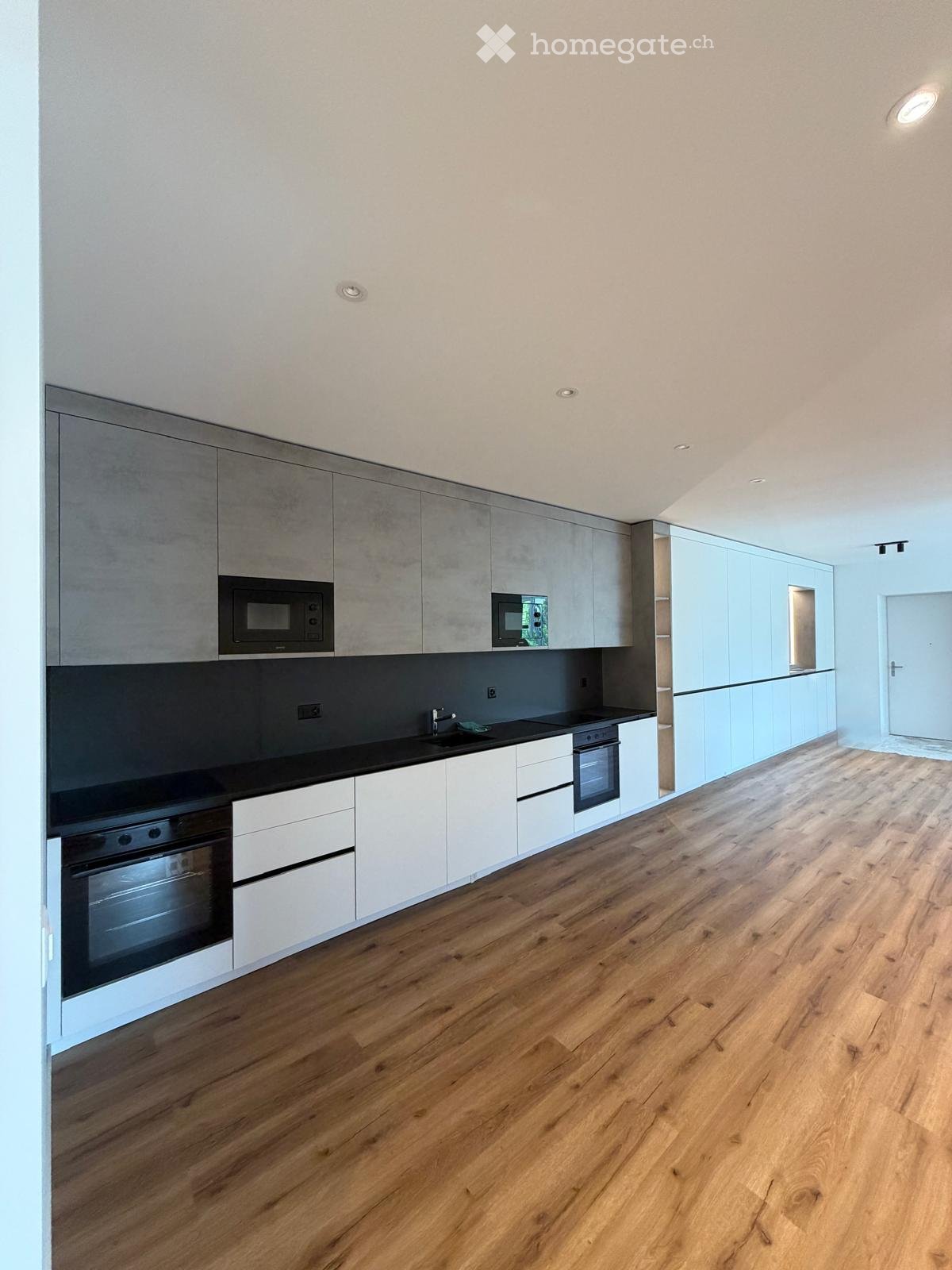 Modern kitchen with light grey and white cabinets, black countertops, and wooden flooring. Features include built-in microwave, oven, and sink.