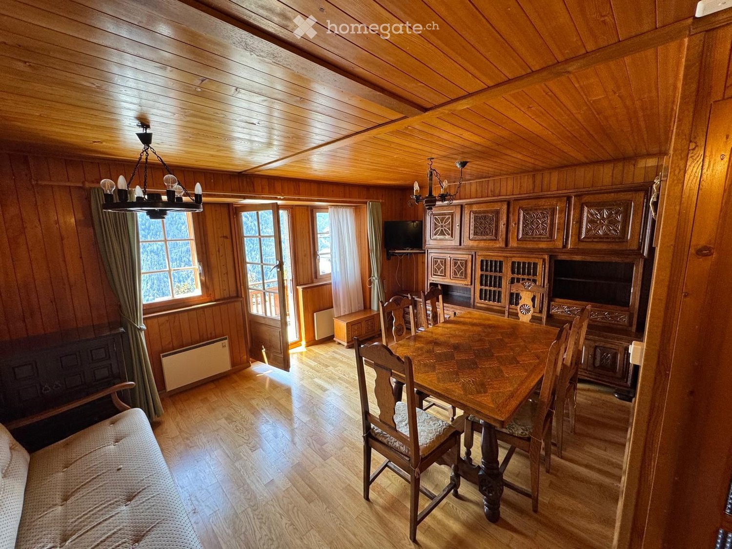 Cozy wooden-paneled room with a dining area, sofa, and cabinetry, featuring large windows and a chandelier.