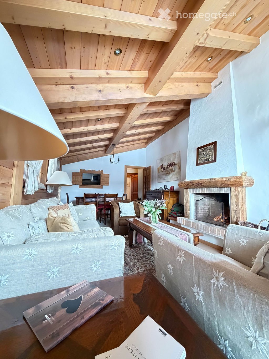 Cozy living room with exposed wooden beams, a fireplace, a sofa, armchairs, coffee table, and dining area visible in the background.