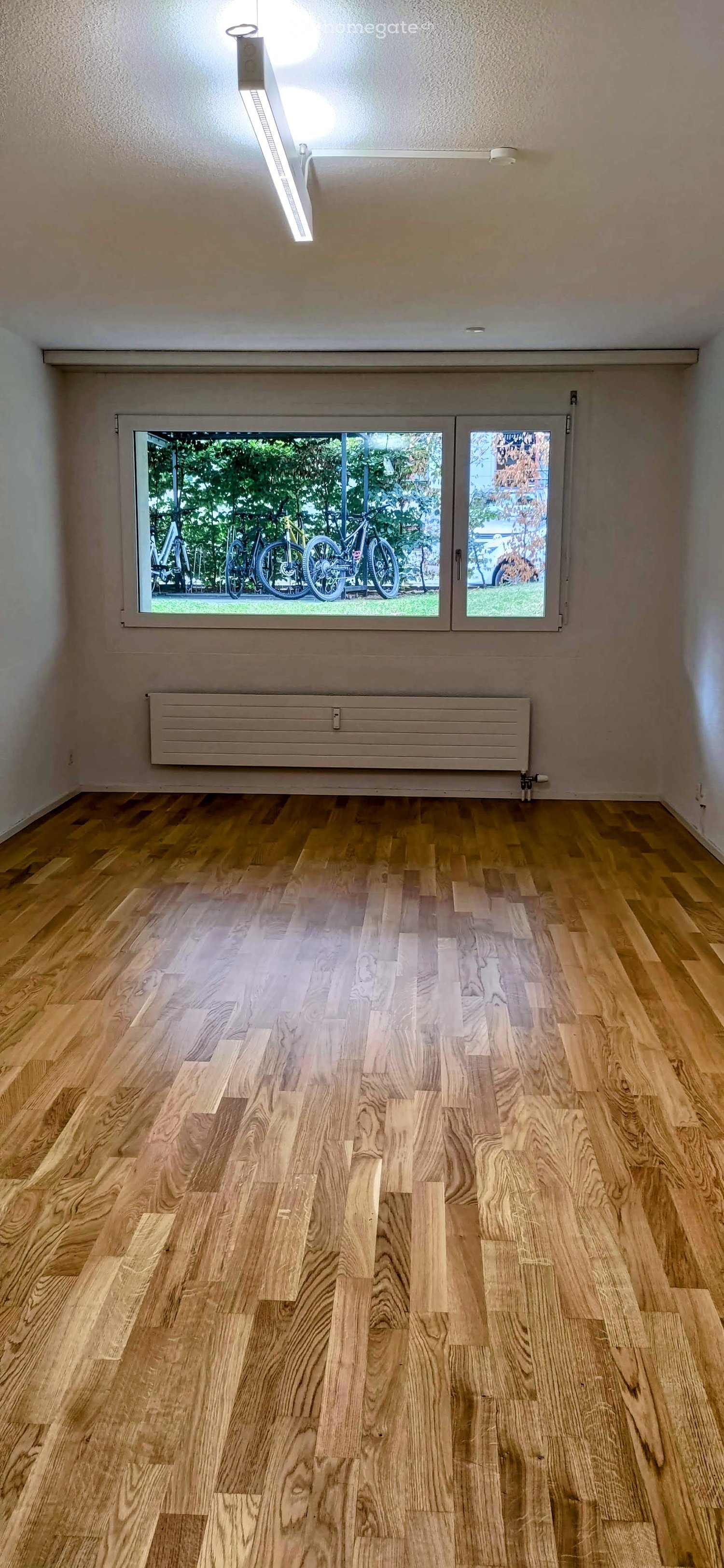 Empty room with wooden flooring, large window showing bicycles outside, radiator, and modern lighting fixture.