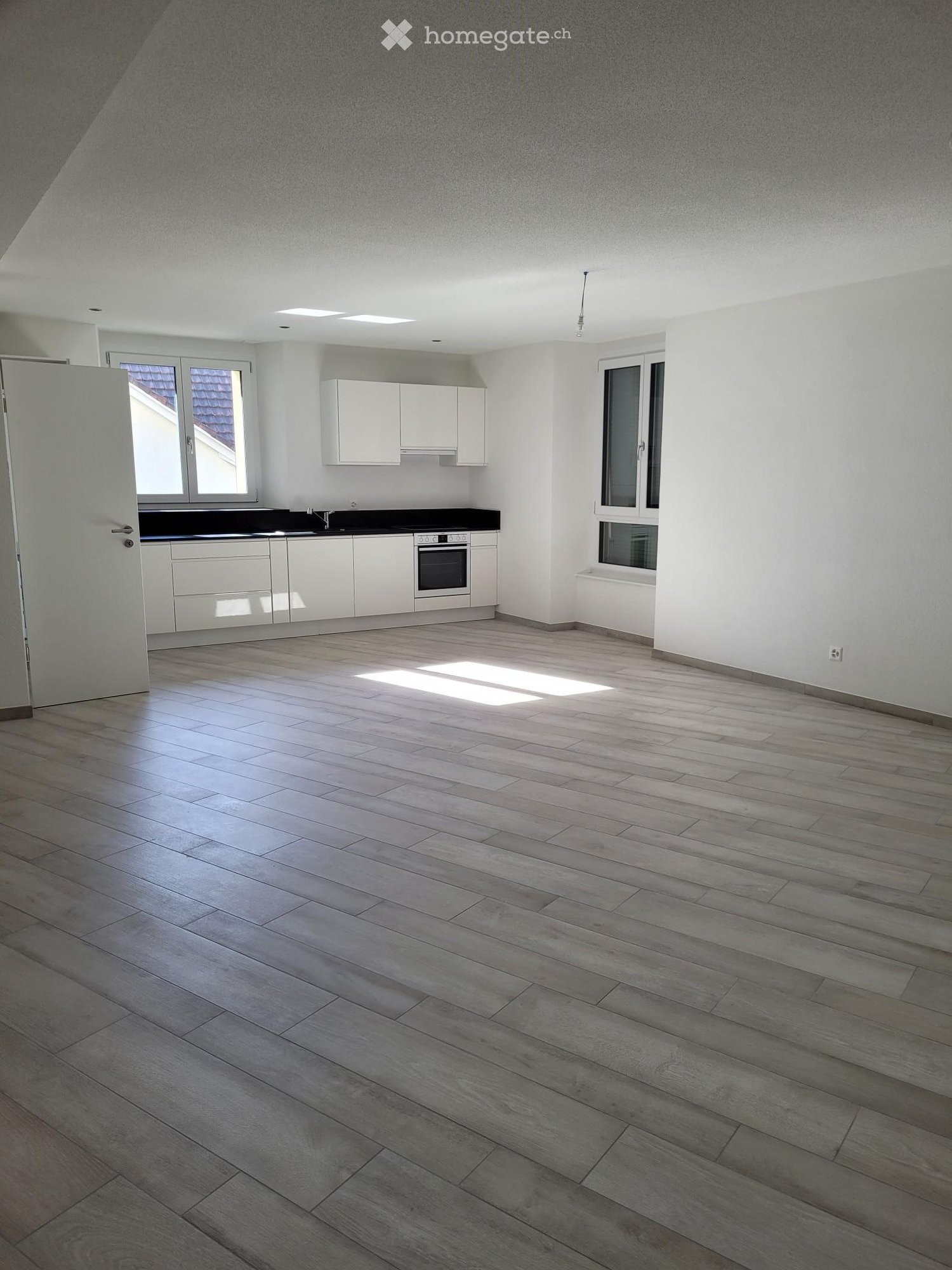 Empty room with light wooden flooring, white walls, and a small kitchen area with white cabinets and appliances visible in the background.