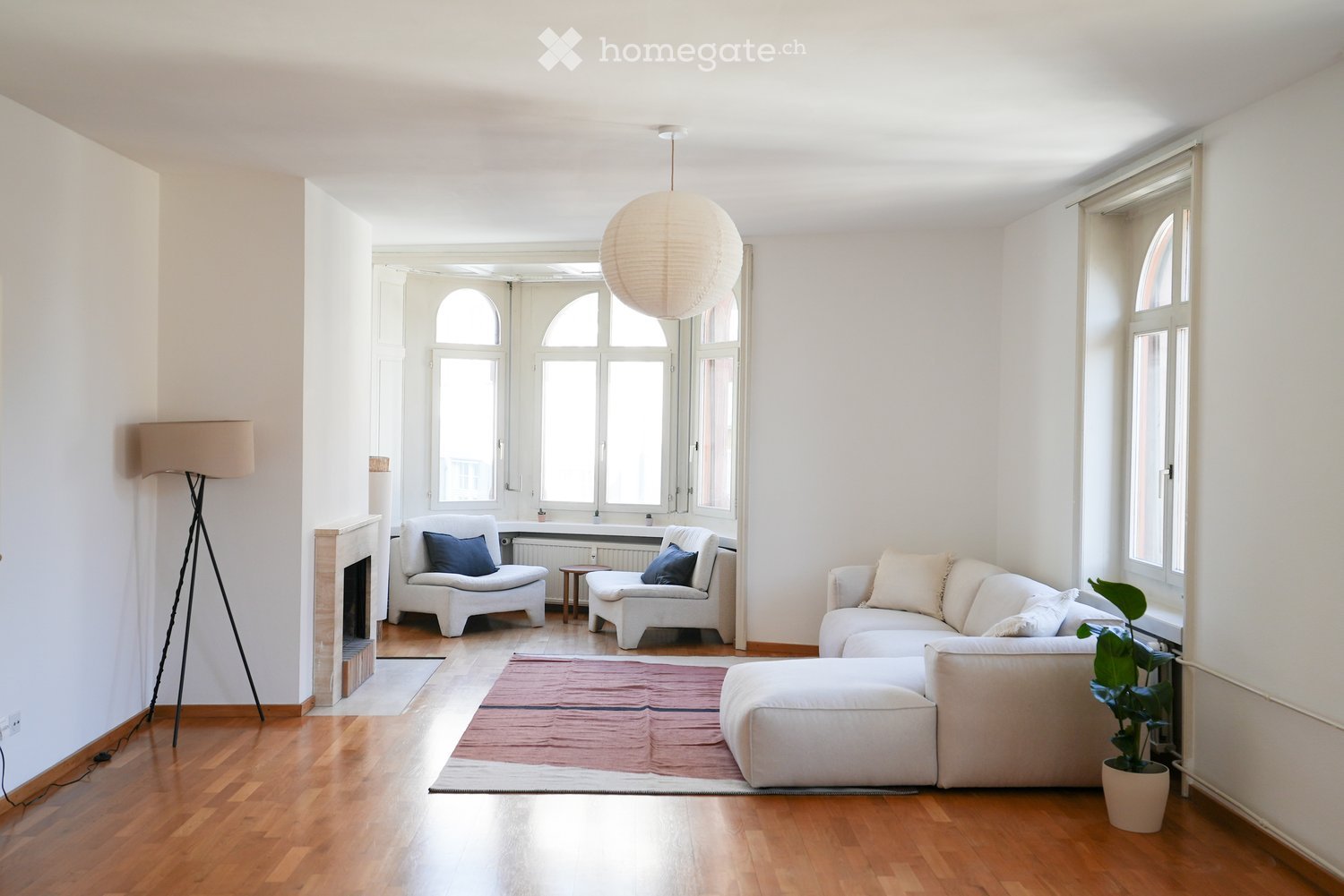 Bright living room with white walls, wooden flooring, large windows, a fireplace, a white sectional sofa, two white armchairs, a small wooden table, a floor lamp, a pendant lamp, and a potted plant.